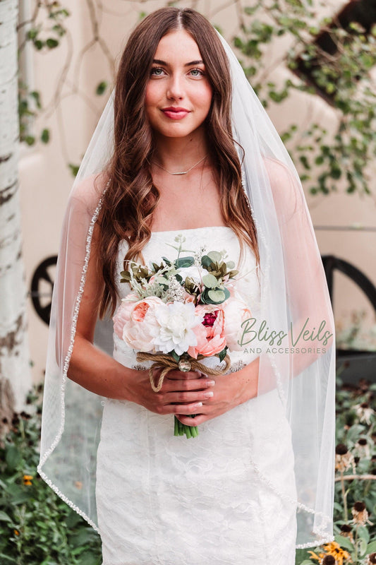 Bride is wearing a one-layer fingertip length beaded wedding veil with pearls in soft white or ivory, made from lightweight sheer tulle for an elegant bridal look.
