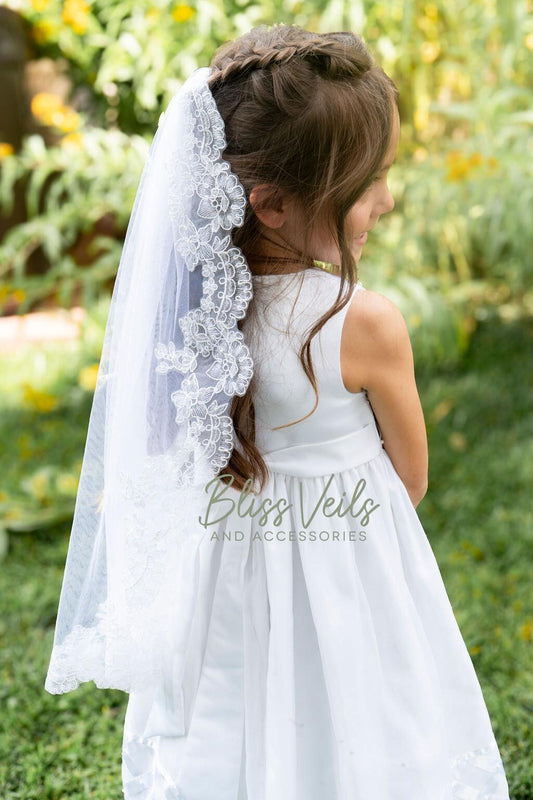 May include: A white communion veil with a lace trim. The veil is made of soft tulle and has a delicate floral design. The veil is worn by a young girl with brown hair. The text on the image reads: 'Bliss Veils and Accessories'.